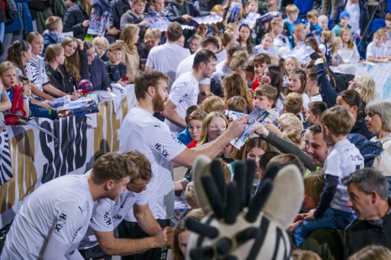 Sportler in weißen Trikots geben Autogramme und unterhalten sich mit einer großen Menge lächelnder Fans, darunter auch Kinder, bei einer Hallenveranstaltung. Die Leute halten Poster und Fanartikel in der Hand, und im Vordergrund ist ein Zebra-Maskottchen zu sehen.