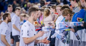 Ein männlicher Sportler in einem weißen Trikot gibt einer Gruppe begeisterter Fans hinter einer Absperrung bei einer Sportveranstaltung Autogramme. Die Fans halten ihm Gegenstände und Erinnerungsstücke hin, die er signieren soll.