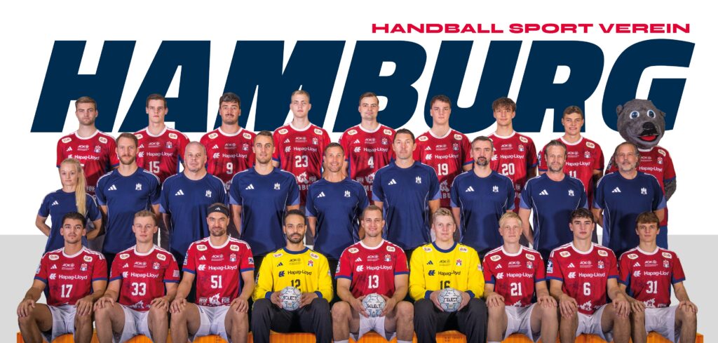 Eine Handballmannschaft in roten und blauen Trikots posiert für ein Gruppenfoto vor einem Schild des Handball Sport Vereins Hamburg. Rechts stehen zwei Torhüter in Gelb und ein Maskottchen in einem Bärenkostüm.