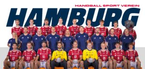 Eine Handballmannschaft in roten und blauen Trikots posiert für ein Gruppenfoto vor einem Schild des Handball Sport Vereins Hamburg. Rechts stehen zwei Torhüter in Gelb und ein Maskottchen in einem Bärenkostüm.