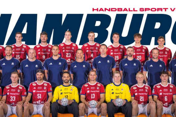 Eine Handballmannschaft in roten und blauen Trikots posiert für ein Gruppenfoto vor einem Schild des Handball Sport Vereins Hamburg. Rechts stehen zwei Torhüter in Gelb und ein Maskottchen in einem Bärenkostüm.