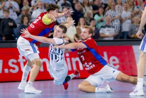 Ein Handballspieler in einem weiß-schwarzen Trikot versucht im Fallen zu schießen, während zwei Gegner in roten Trikots versuchen, ihn während eines intensiven Spiels zu blocken, mit einer jubelnden Menge im Hintergrund.