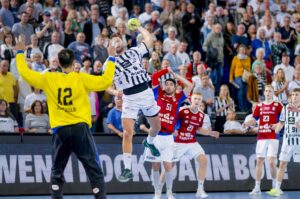 Ein Handballspieler in einem weiß-schwarzen Trikot springt, um den Ball in Richtung Tor zu werfen, während ein Torwart in Gelb und gegnerische Spieler in roten Trikots verteidigen. Im Hintergrund schaut eine große Menschenmenge aufmerksam zu.