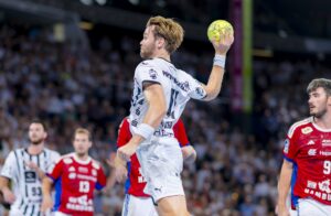 Ein männlicher Handballspieler in einem weißen Trikot springt und bereitet sich darauf vor, während eines Spiels einen gelben Ball zu werfen, während andere Spieler in roten und weißen Trikots und eine verschwommene Menschenmenge im Hintergrund zu sehen sind.