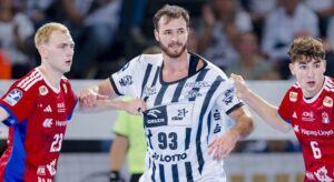 Drei männliche Handballspieler sind während eines Spiels in Aktion. Der Spieler in der Mitte, der ein weißes Trikot trägt, wird von zwei Spielern in roten Trikots verteidigt, von denen sich einer das weiße Trikot schnappt.