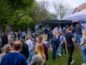 Eine große Gruppe von Menschen trifft sich im Freien bei einem zwanglosen Treffen in der Nähe eines modernen Gebäudes, das von Bäumen und Grünflächen umgeben ist. Einige stehen in kleinen Gruppen und unterhalten sich, während andere sitzen oder herumlaufen.