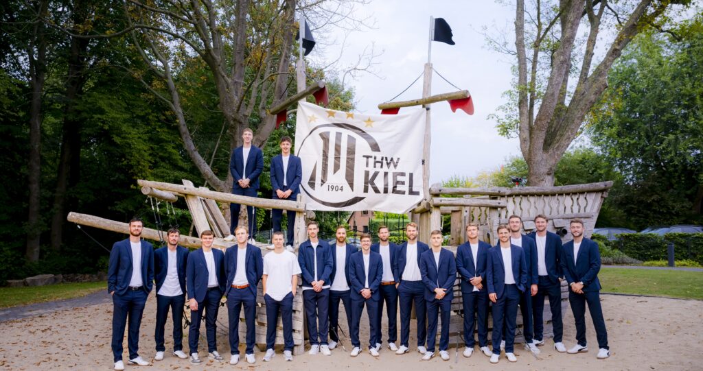 Eine Gruppe von Männern in passenden blauen Anzügen und weißen Turnschuhen steht und sitzt auf einer großen Holzkonstruktion im Freien, hinter ihnen hängt ein Banner des THW Kiel zwischen Bäumen.