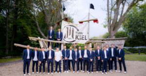 Eine Gruppe von Männern in passenden blauen Anzügen und weißen Turnschuhen steht und sitzt auf einer großen Holzkonstruktion im Freien, hinter ihnen hängt ein Banner des THW Kiel zwischen Bäumen.