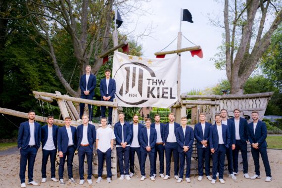 Eine Gruppe von Männern in passenden blauen Anzügen und weißen Turnschuhen steht und sitzt auf einer großen Holzkonstruktion im Freien, hinter ihnen hängt ein Banner des THW Kiel zwischen Bäumen.