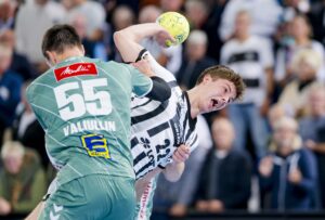 Ein Handballspieler in einem schwarz-weiß gestreiften Trikot springt zum Wurf, während er von einem Spieler in einem grünen Trikot mit dem Namen VAILULIN und der Nummer 55 verteidigt wird. Im unscharfen Hintergrund ist eine Menschenmenge zu sehen.