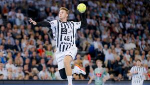 Ein männlicher Handballspieler in einem schwarz-weiß gestreiften Trikot springt in der Luft, um während eines Spiels einen gelben Ball zu werfen, mit einem unscharfen, überfüllten Stadionpublikum im Hintergrund.