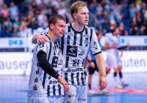 Zwei männliche Handballspieler in schwarz-weiß gestreiften Trikots stehen nebeneinander auf einem Spielfeld, einer hat den Arm um den anderen gelegt. Beide wirken konzentriert, im Hintergrund ist eine verschwommene Menschenmenge zu sehen.