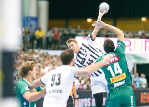 Ein Handballspiel der Männer in Aktion, bei dem ein Spieler im weißen Trikot springt, um den Ball zu schießen, während er von einem Spieler im grünen Trikot geblockt wird. Im Hintergrund sind andere Spieler und eine Zuschauermenge zu sehen.