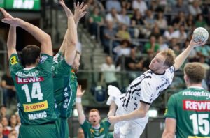 Ein Handballspieler in einem weißen Trikot springt, um den Ball zu schießen, während drei Verteidiger in grünen Trikots ihn blockieren. Die Aktion findet in einer Halle statt, mit einer unscharfen Menschenmenge im Hintergrund.