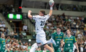 Ein Handballspieler in einem weißen Trikot springt in die Luft, um den Ball zu werfen, während drei gegnerische Spieler in grünen Trikots ihn während eines Spiels in einer mit Zuschauern gefüllten Halle genau beobachten.