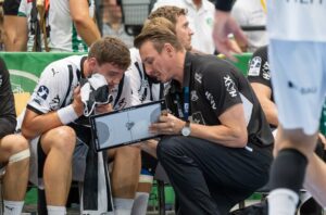 Ein Trainer kniet neben einem sitzenden Handballspieler und bespricht mit einem Klemmbrett die Strategie. Der Spieler hört aufmerksam zu und hält sich ein Handtuch vor das Gesicht, während Mannschaftskameraden und Betreuer in der Nähe sind.