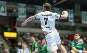 Ein Handballspieler in einem weißen Trikot mit der Nummer 7 wirft den Ball während eines Spiels. Im Hintergrund sind Spieler in grünen Trikots und Schiedsrichter zu sehen, sowie ein verschwommenes Publikum und Banner.