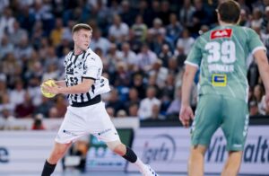 Ein männlicher Handballspieler in einem schwarz-weiß gestreiften Trikot bereitet sich darauf vor, den Ball zu werfen, während ein anderer Spieler in einem grünen Trikot ihm auf einem Spielfeld gegenübersteht.
