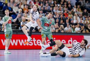 Zwei Handballspieler in weißen und schwarzen Trikots stoßen während eines intensiven Spiels mit zwei Spielern in grünen Trikots zusammen. Ein Spieler in weißer Uniform ist in der Luft, während ein anderer am Boden liegt. Die Zuschauer im Hintergrund schauen zu.