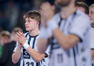 Ein junger männlicher Sportler in einem schwarz-weiß gestreiften Trikot steht mit verschränkten Händen da und blickt nach vorne. Andere Mannschaftskameraden in ähnlichen Trikots sind unscharf im Vorder- und Hintergrund zu erkennen. Die Atmosphäre wirkt ernst oder nachdenklich.