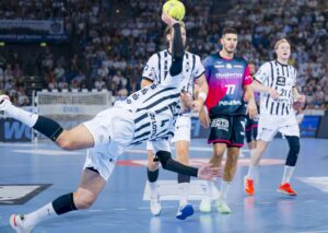 Ein Handballspieler in einem weiß gestreiften Trikot springt und wirft den Ball während eines Spiels in die Luft, während drei gegnerische Spieler zuschauen und eine Menschenmenge im Hintergrund zu sehen ist.