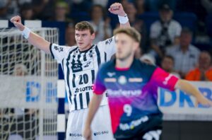 Ein Handballspieler in einem schwarz-weiß gestreiften Trikot reißt in der Nähe des Tores jubelnd beide Arme hoch, während ein anderer Spieler in einem dunklen Trikot mit rosa Ärmeln im Vordergrund steht. Im Hintergrund sieht eine Menschenmenge zu.