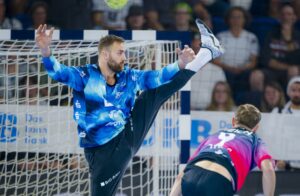 Ein Handballtorwart streckt sein Bein hoch, um einen Schuss abzublocken, während ein gegnerischer Spieler mit der Trikotnummer 5 in der Nähe läuft. Im Hintergrund sehen Zuschauer zu.