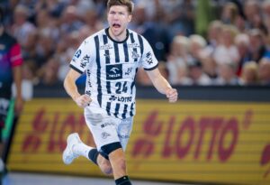 Ein männlicher Handballspieler in einem schwarz-weiß gestreiften Trikot rennt und jubelt während eines Spiels, während im Hintergrund eine Menschenmenge und eine Lotto-Werbung zu sehen sind.