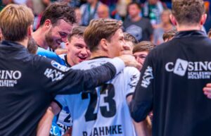 Eine Gruppe von männlichen Sportlern in Sporttrikots sitzt zusammen, lächelt und feiert. Auf dem Rücken eines Spielers steht der Name JAHMKE und die Nummer 23. Im Hintergrund ist eine unscharfe Menschenmenge zu sehen.