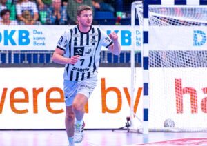 Ein männlicher Handballspieler in einem weiß-schwarz gestreiften Trikot mit der Nummer 7 läuft mit geballter Faust über einen Hallenplatz. Im Hintergrund sind ein Tor, Werbetafeln und Zuschauer auf der Tribüne zu sehen.