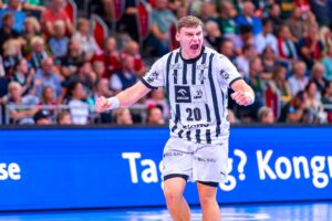 Ein männlicher Handballspieler in einem weiß-schwarz gestreiften Trikot mit der Nummer 20 jubelt leidenschaftlich auf dem Spielfeld. Eine Zuschauermenge füllt den Hintergrund.