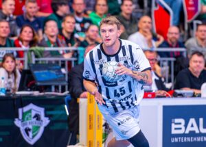 Ein Handballspieler in einem weiß-schwarzen Trikot bereitet sich während eines Spiels auf den Wurf vor, während das Publikum auf der Tribüne hinter ihm zusieht.