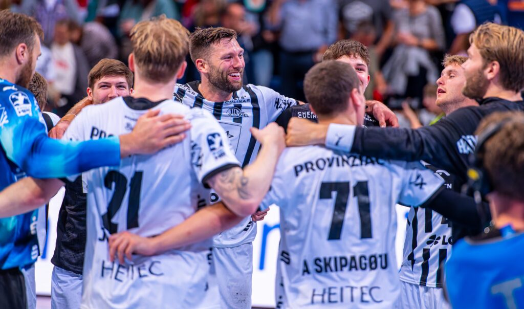 Eine Gruppe männlicher Handballspieler in weißen und schwarzen Trikots steht in einem Kreis, lächelt und feiert mit umschlungenen Armen, während im Hintergrund ein verschwommenes Publikum zuschaut.