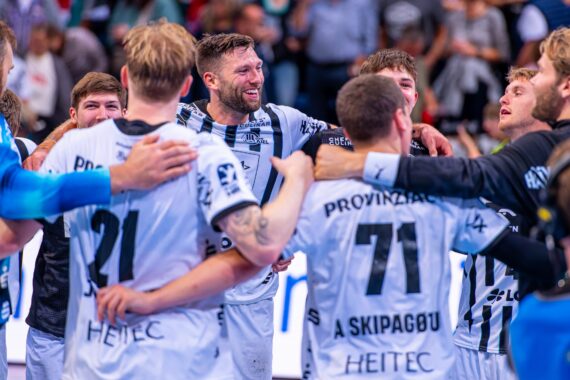Eine Gruppe männlicher Handballspieler in weißen und schwarzen Trikots steht in einem Kreis, lächelt und feiert mit umschlungenen Armen, während im Hintergrund ein verschwommenes Publikum zuschaut.