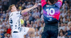 Ein Handballspieler in einem weißen Trikot springt und bereitet sich darauf vor, den Ball in Richtung Tor zu werfen, während ein Torwart in einem schwarz-rosafarbenen Trikot mit der Nummer 10 versucht, den Schuss zu blocken. Eine Menschenmenge schaut im Hintergrund zu.