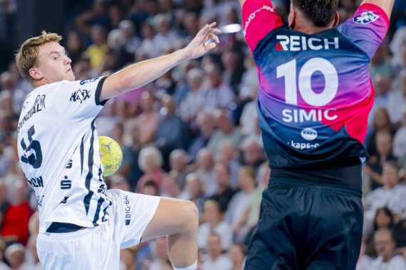 Ein Handballspieler in einem weißen Trikot springt und bereitet sich darauf vor, den Ball in Richtung Tor zu werfen, während ein Torwart in einem schwarz-rosafarbenen Trikot mit der Nummer 10 versucht, den Schuss zu blocken. Eine Menschenmenge schaut im Hintergrund zu.