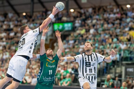Drei männliche Handballspieler, zwei in weiß gestreiften Trikots und einer in grün, springen und greifen nach dem Ball in einer mit Zuschauern gefüllten Halle. Im Hintergrund sind die Zuschauer und helle Bildschirme zu sehen.