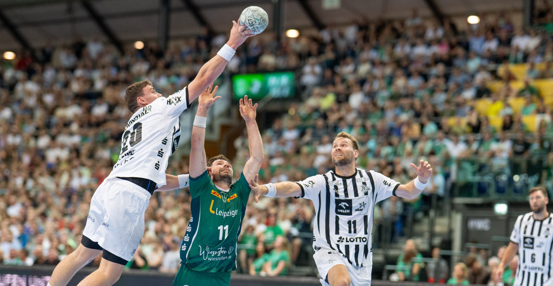 Drei männliche Handballspieler, zwei in weiß gestreiften Trikots und einer in grün, springen und greifen nach dem Ball in einer mit Zuschauern gefüllten Halle. Im Hintergrund sind die Zuschauer und helle Bildschirme zu sehen.