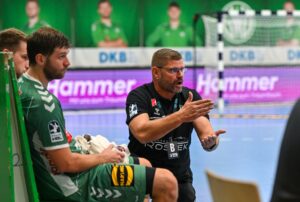 Ein Handballtrainer gibt einem sitzenden Spieler in einem grünen Trikot während eines Spiels leidenschaftlich Anweisungen, während im Hintergrund andere Spieler und ein Tor zu sehen sind.