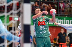 Ein männlicher Handballspieler in einem grünen Trikot bereitet sich darauf vor, den Ball während eines Spiels zu werfen, während im Hintergrund Gegner und ein Tor zu sehen sind. Die Zuschauer schauen von der Tribüne aus zu.