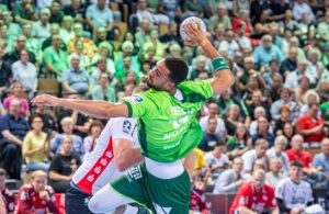 Ein Handballspieler in einem grünen Trikot springt mit dem Ball, um ein Tor zu erzielen, während ein Verteidiger in einem weiß-roten Trikot versucht, ihn abzuwehren. Ein dicht gedrängtes Publikum verfolgt das intensive Spiel.