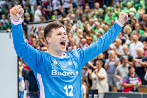 Ein Handballspieler, der ein blaues Trikot mit der Nummer 12 trägt, jubelt mit erhobenen Fäusten. Die Menge im Hintergrund scheint begeistert und jubelt.