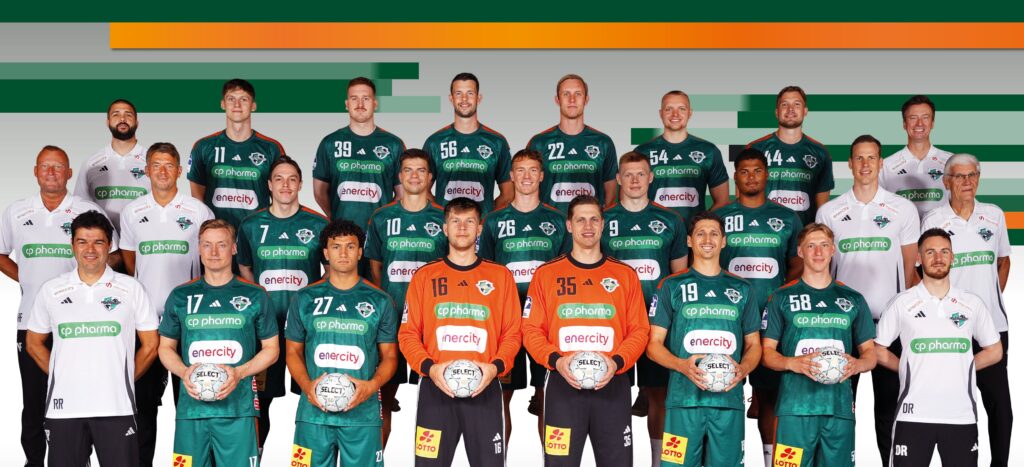 25_vor_Hannover_Team Eine Handballmannschaft der Männer posiert für ein Gruppenfoto in grünen Trikots mit Mitarbeitern in weißen Hemden, die Handbälle halten, vor einem Hintergrund mit grünen und orangefarbenen Streifen.