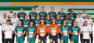 Eine Handballmannschaft der Männer posiert für ein Gruppenfoto in grünen Trikots mit Mitarbeitern in weißen Hemden, die Handbälle halten, vor einem Hintergrund mit grünen und orangefarbenen Streifen.
