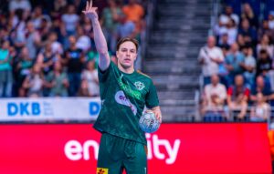 Ein männlicher Handballspieler in einem grünen Trikot hält einen Ball in einer Hand und hebt die andere Hand, um während eines Spiels ein Zeichen zu geben, mit einer verschwommenen Menschenmenge und roter und blauer Werbung im Hintergrund.