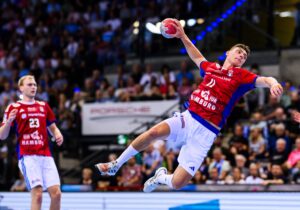 Ein männlicher Handballspieler in einem rot-blauen Trikot springt in die Luft und bereitet sich auf den Wurf vor, während ein anderer Spieler im Hintergrund zusieht. Die Szene spielt sich in einer überfüllten Halle ab.