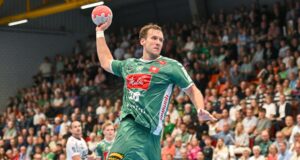 Ein männlicher Handballspieler in einem grünen Trikot springt mit erhobenem Arm, um den Ball zu werfen. Im Hintergrund füllt sich die Tribüne mit einer Menge von Zuschauern.
