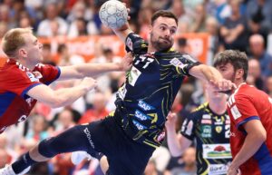 Ein Handballspieler in einem marineblauen und gelben Trikot springt in der Luft und bereitet sich auf den Wurf vor, während zwei Gegner in Rot und Blau versuchen, ihn während eines Spiels zu blockieren. Im Hintergrund ist eine unscharfe Menschenmenge zu sehen.