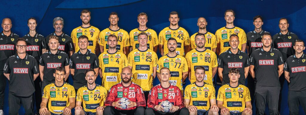 Eine Herren-Handballmannschaft posiert für ein Gruppenfoto. Die Spieler tragen gelbe Trikots, zwei von ihnen haben rote Torwarttrikots an und halten Bälle. Die Trainer und Betreuer in schwarzen Hemden stehen hinter und neben ihnen, alle schauen in die Kamera und lächeln.