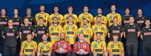 Eine Herren-Handballmannschaft posiert für ein Gruppenfoto. Die Spieler tragen gelbe Trikots, zwei von ihnen haben rote Torwarttrikots an und halten Bälle. Die Trainer und Betreuer in schwarzen Hemden stehen hinter und neben ihnen, alle schauen in die Kamera und lächeln.